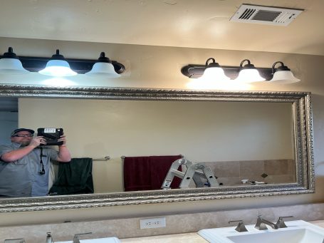 A bathroom mirror with lights above, reflecting a person and a ladder.