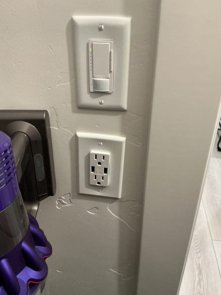 Light switch and electrical outlet on a wall near a vacuum cleaner.